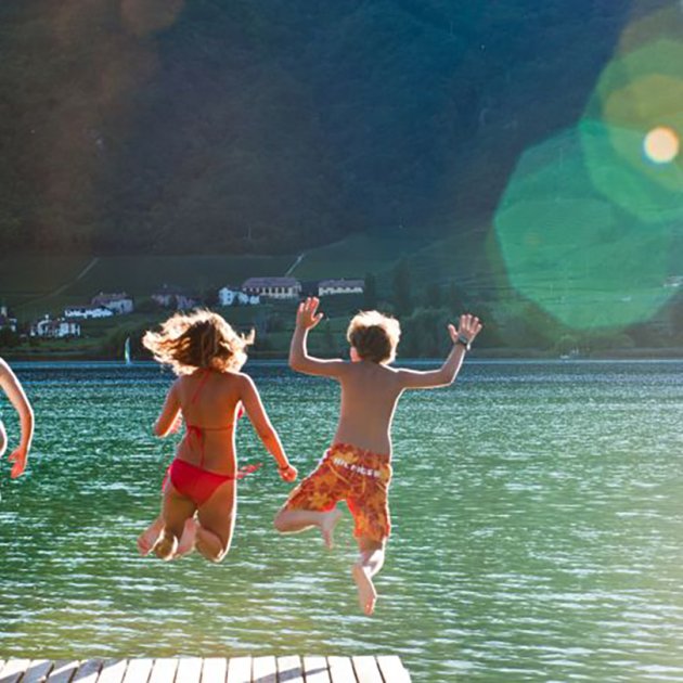 Kinder springen lachend von einem Steg in einen klaren See
