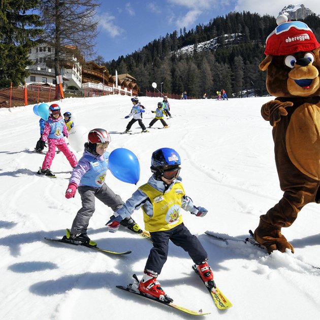 Maskottchen-Bär fährt auf Schnee mit Kindern, die Skifahren lernen, und bunten Luftballons.
