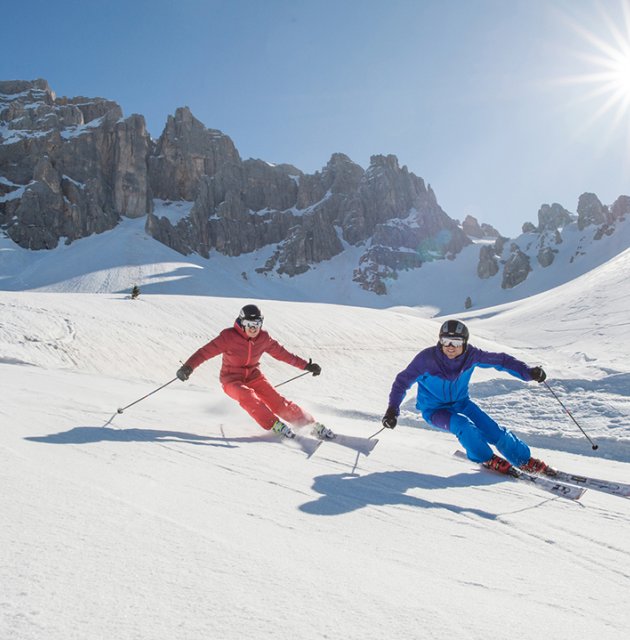 Zwei Skifahrer in roten und blauen Anzügen fahren eine verschneite Piste mit felsigen Bergen und strahlender Sonne hinunter.